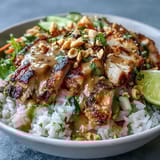 Tender grilled chicken slices rest on coconut rice with crisp cabbage, carrots, and cucumber, finished with a luscious peanut drizzle and lime wedges.