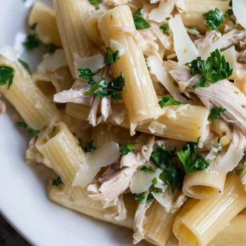 A large skillet holds creamy Roasted Garlic Chicken Penne, garnished with fresh parsley and extra Parmesan, steaming on a rustic table. 