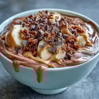 A close-up of a Chocolate Peanut Butter Smoothie Bowl in a white bowl, topped with banana slices, granola, chopped dark chocolate, and a peanut butter drizzle.